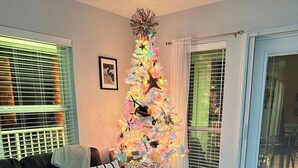 A living room with a colorful Christmas tree, a sofa with cushions, a coffee table with decorative items, and a window with blinds. - Beautiful views of beach, sunset, sunrise and the Blue Herons at the pond... (Pensacola)