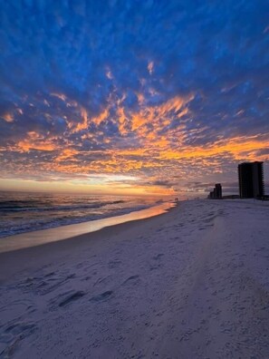 Ein Strand bei Sonnenuntergang, mit Wellen und einigen Gebäuden in der Ferne.