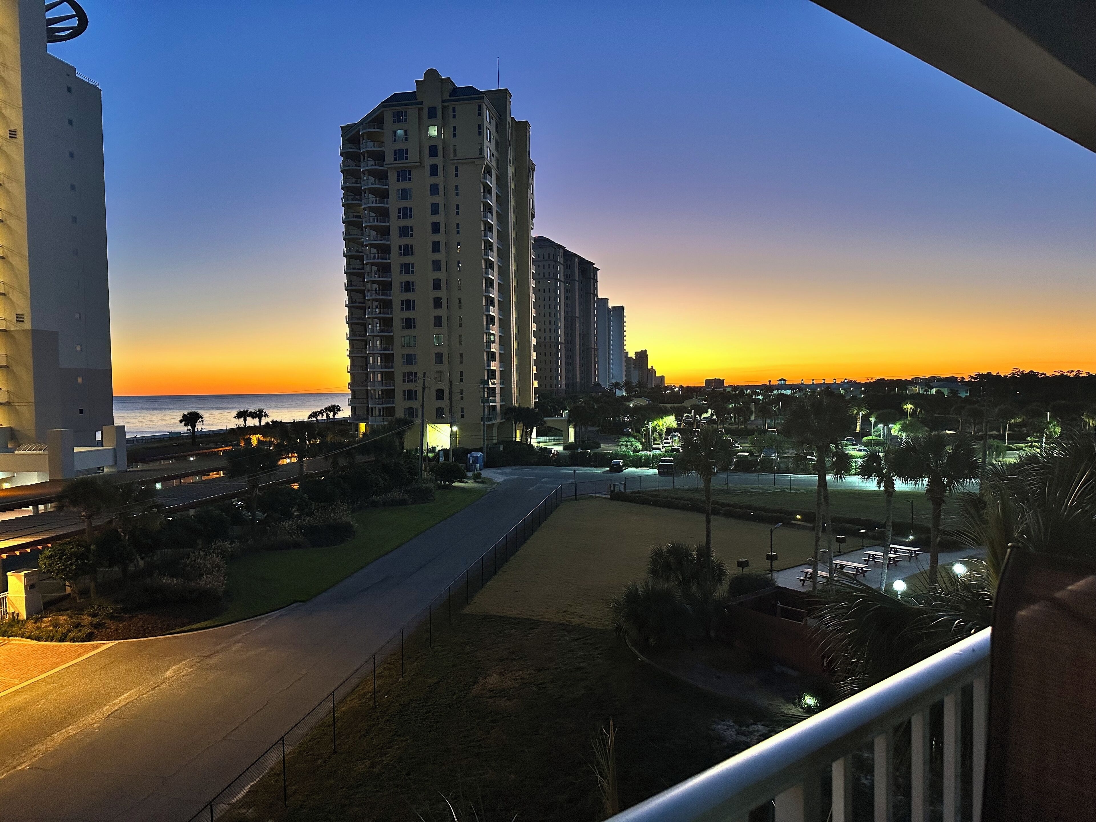 A high-rise building with multiple balconies, a road in the foreground, and a sunset in the background.
