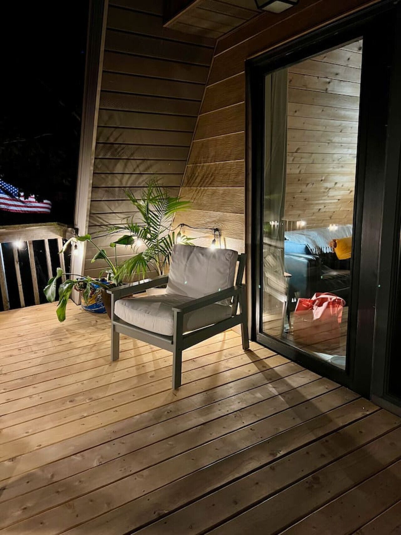 A wooden deck with a chair and potted plants, a sliding glass door leading to a room with a bed and a suitcase.