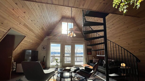 A cozy loft bedroom with a sloped ceiling, a spiral staircase, a large window with a view, and a modern living area with a sofa, chairs, and a coffee table. - Waterfront A-Frame on the Marina • Lake Pepin (Pepin)