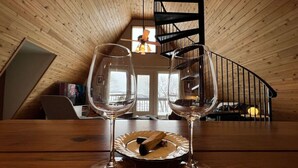 A wooden dining table with two wine glasses and a plate with a wooden object. A spiral staircase and a window with a view of trees are visible in the background. - Waterfront A-Frame on the Marina • Lake Pepin (Pepin)