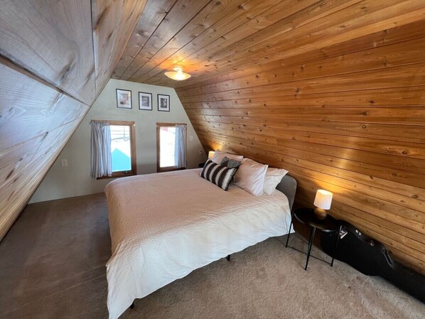 A bedroom with a sloped ceiling, a bed with white bedding, two windows with curtains, and a guitar on a stand.