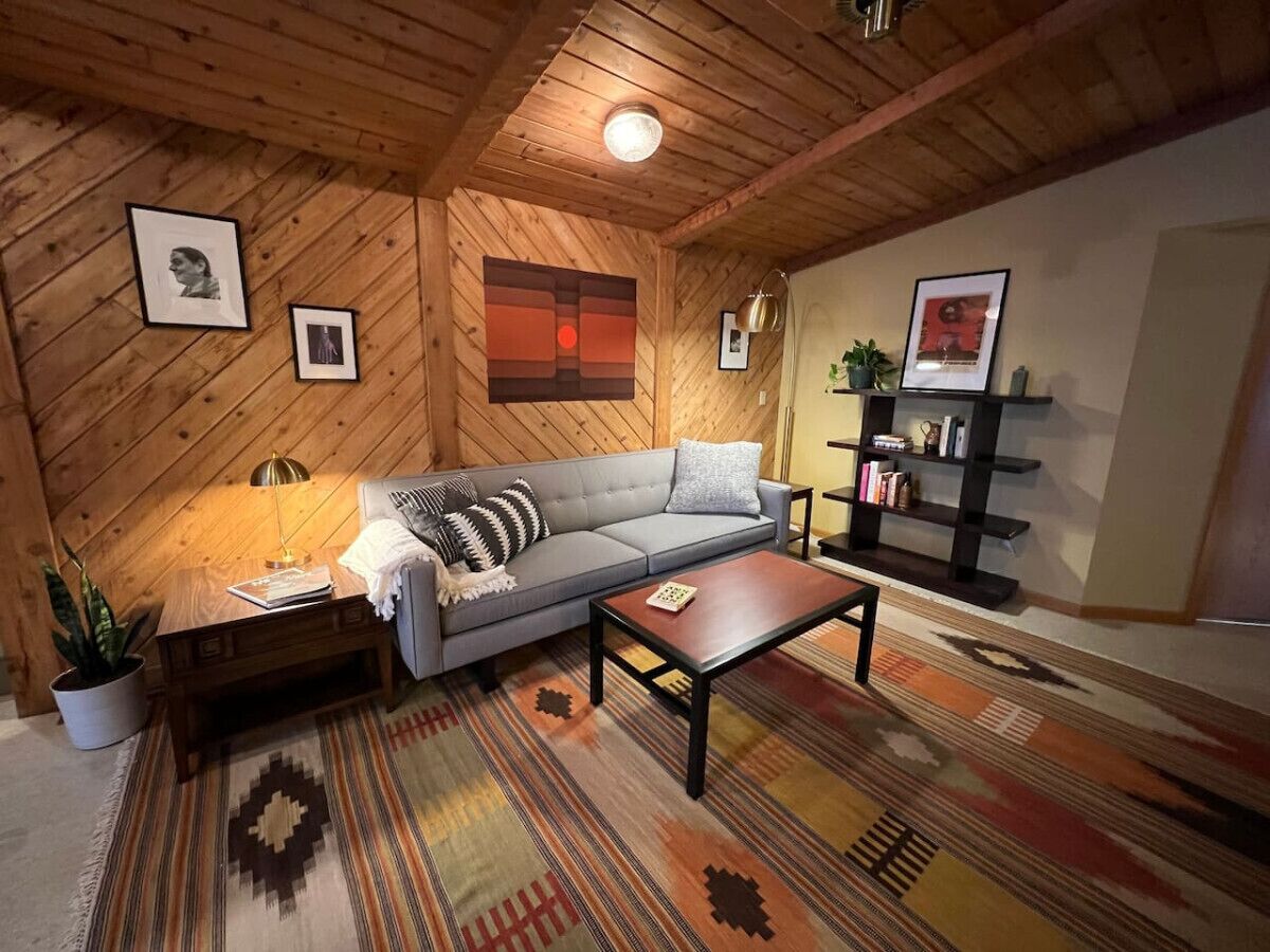 Ein gemütlicher Raum mit Holzboden, einem grauen Sofa, einem Holztisch und einem gemusterten Teppich.