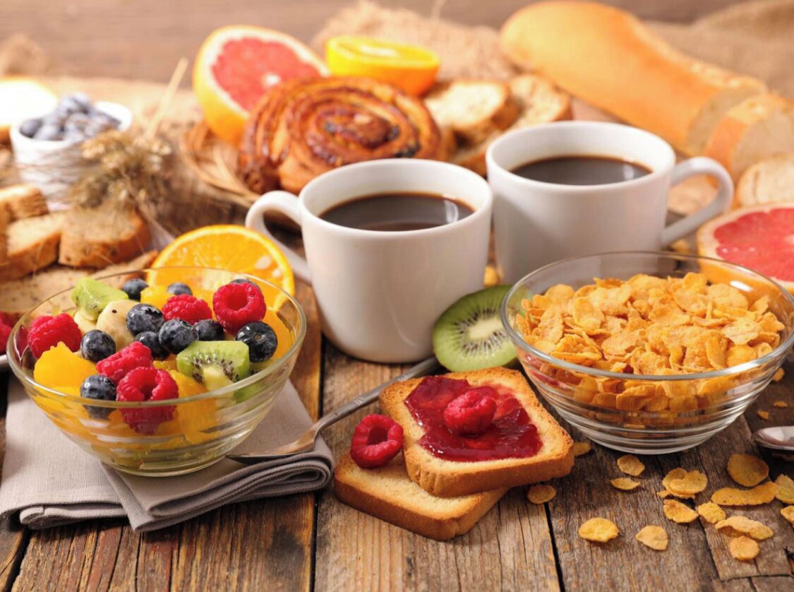 Une table en bois dressée pour le petit-déjeuner, avec des toasts, des céréales, des fruits et des tasses de café.