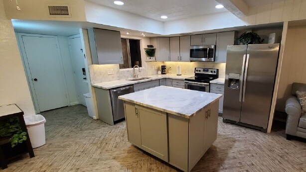 Een moderne keuken met een centraal eiland, roestvrijstalen apparatuur en een grijze bank in de woonkamer.