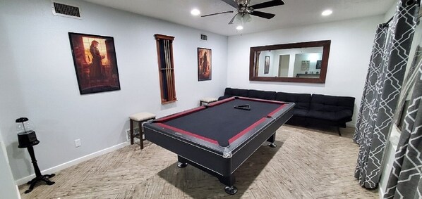 Une table de billard dans une pièce dotée d’un ventilateur de plafond, d’un canapé et d’œuvres d’art encadrées accrochées aux murs.