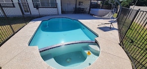 Une piscine avec un jacuzzi, entourée d’une clôture et d’un espace patio.