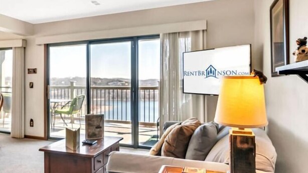 A hotel room with a large sliding glass door leading to a balcony with a view of the landscape. There is a sofa, a lamp, and a framed picture on the wall. - Cozy Condo with Table Rock Lake Views, Free Tickets & 5 Minutes from Silver Dollar City (Branson)