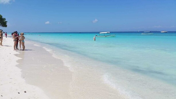 Ein Strand mit klarem türkisfarbenem Wasser, einige Personen im Sand und ein Boot in der Ferne.