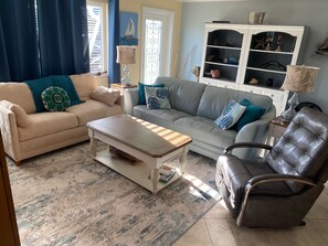 Una sala de estar con un sofá color beige, un sofá gris, una mesa de centro de madera, un sillón de cuero y una estantería blanca con objetos decorativos.