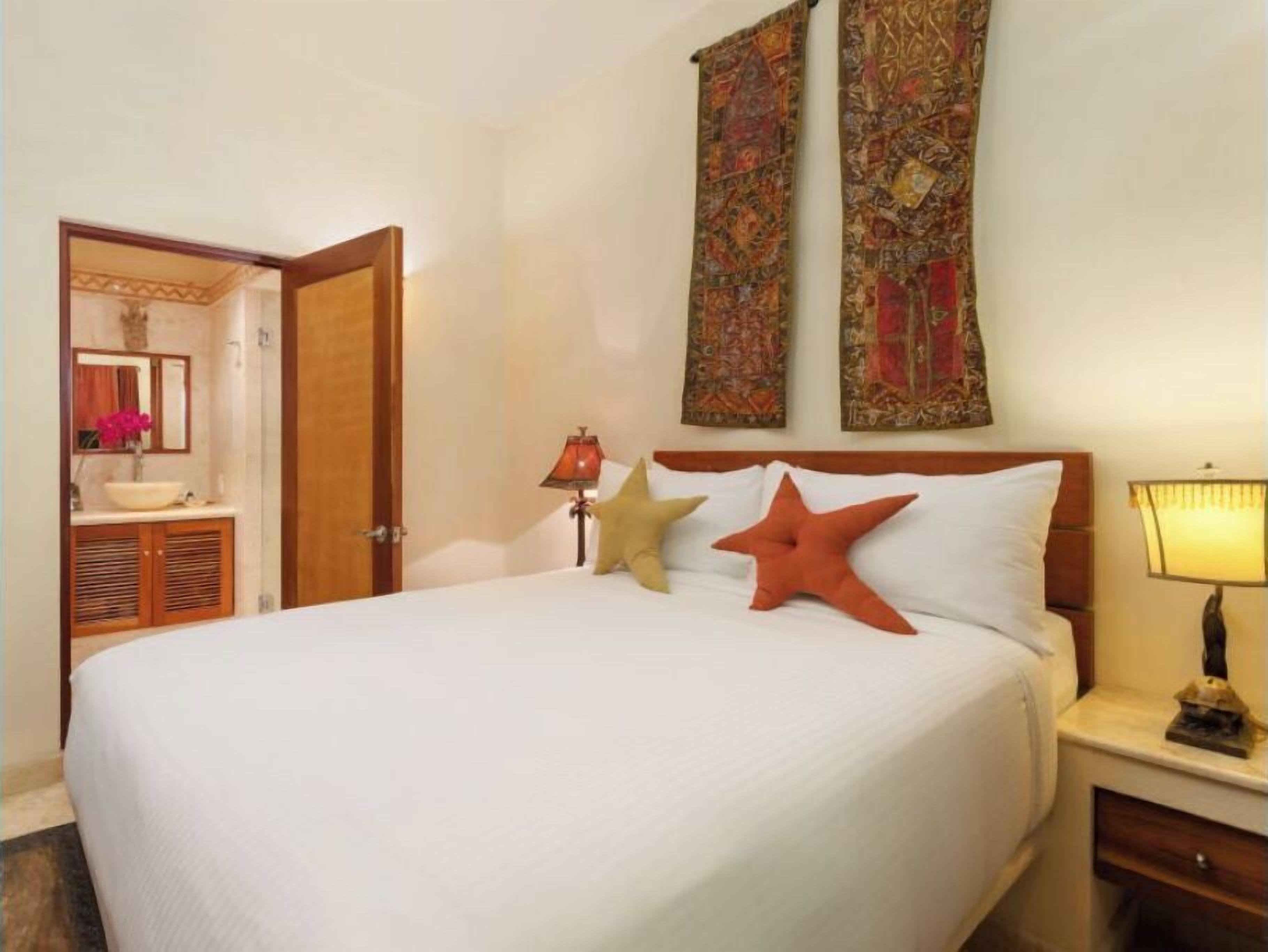 Habitación de hotel con una cama grande, dos almohadones decorativos, una mesita de noche con lámpara, un cabecero de madera y un baño visible a través de una puerta abierta.
