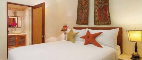Habitación de hotel con una cama grande, dos almohadones decorativos, una mesita de noche con lámpara, un cabecero de madera y un baño visible a través de una puerta abierta.