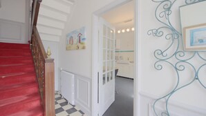 Um corredor com escada revestida de tapete vermelho, radiador branco e porta que dá para um espaço com banheiro e pia.