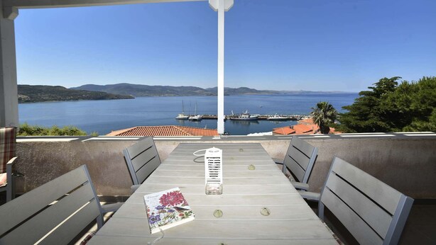 A covered outdoor seating area with a table and chairs, overlooking a body of water with boats and a distant shoreline. - Heliades maisonettes (Lesvos)