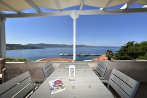 A covered outdoor seating area with a table and chairs, overlooking a body of water with boats and a distant shoreline. - Heliades maisonettes (Lesvos)