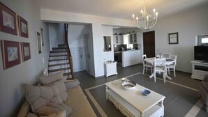 A spacious living room with a sofa, a dining table with chairs, a kitchen area with cabinets, and a staircase leading to another level. - Heliades maisonettes (Lesvos)