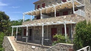 A two-story stone building with a red-tiled roof, a covered outdoor seating area, and a staircase leading to the upper floor. - Heliades maisonettes (Lesvos)