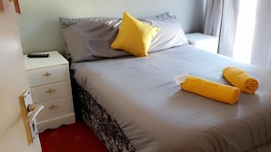 A neatly made bed with a grey duvet and two rolled towels, a white bedside table with a remote control, and a door with a handle. - Bel Reve Hotel (Blackpool)