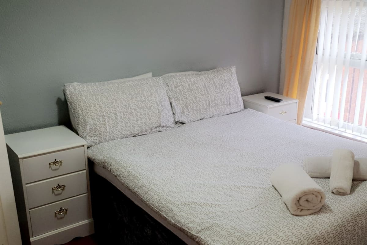 A neatly made bed with a patterned bedspread, two pillows, and two rolled towels. There is a bedside table with a remote control and a drawer unit with three drawers.