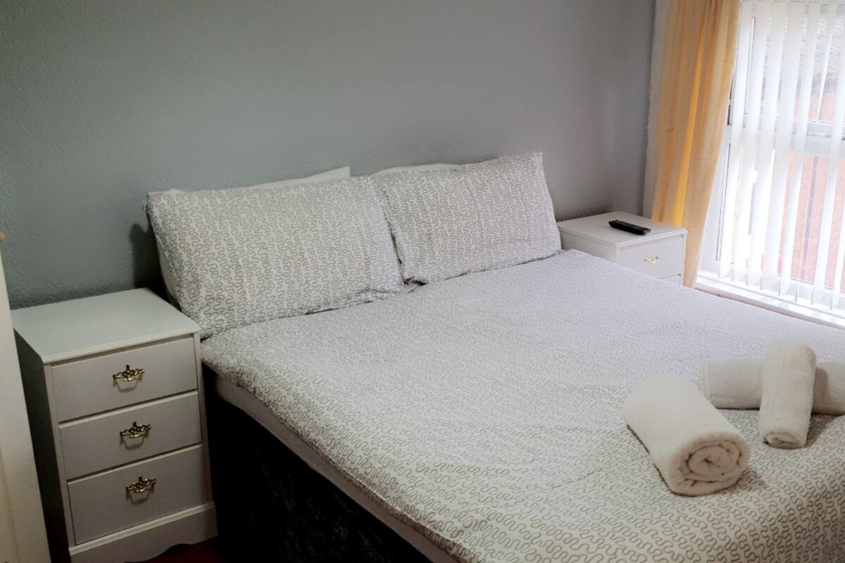 A neatly made bed with a patterned bedspread, two pillows, and two rolled towels. There is a bedside table with a remote control and a drawer unit with three drawers.