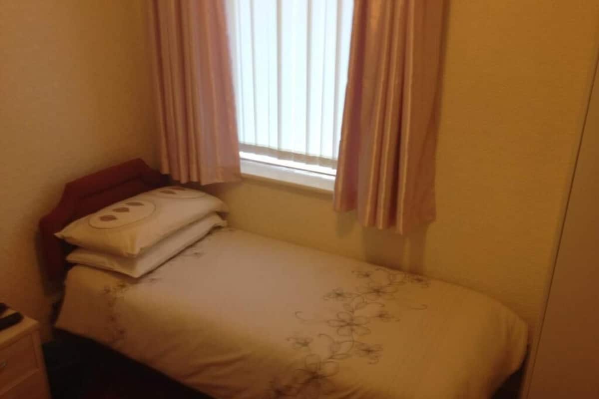 A small, single-bed room with a window, curtains, and a bedside table.