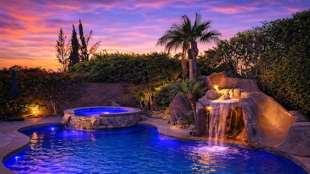 A swimming pool with a hot tub and a waterfall feature, surrounded by lush greenery and a stone wall.