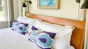 Una cama bien hecha con cabecera de madera, dos almohadas estampadas, un cuadro enmarcado encima de la cama y dos lámparas fijadas a la pared.