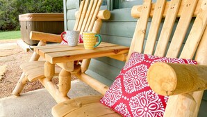 Una silla Adirondack de madera con un cojín estampado en rojo y blanco, una mesa de madera con dos tazas decoradas, y un banco de madera.