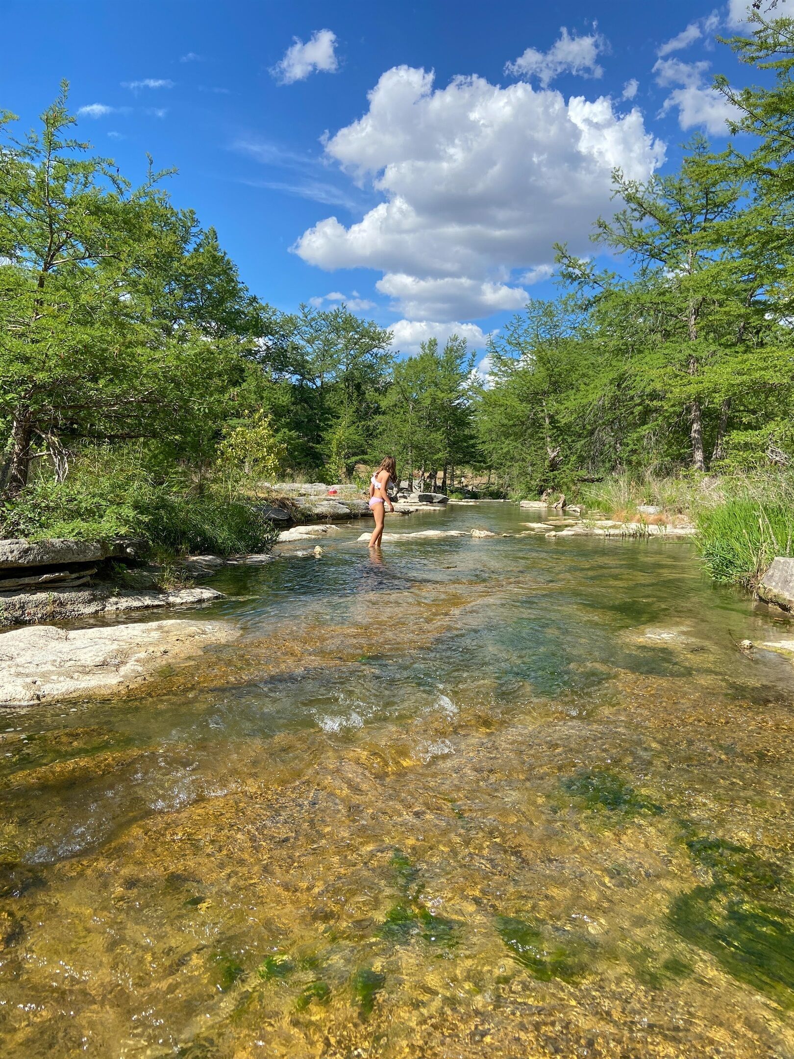 A clear river with rocks and trees on both sides, a person standing in the water, and a partly cloudy sky.