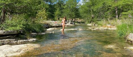 A clear river with rocks and trees on both sides, a person standing in the water, and a partly cloudy sky.