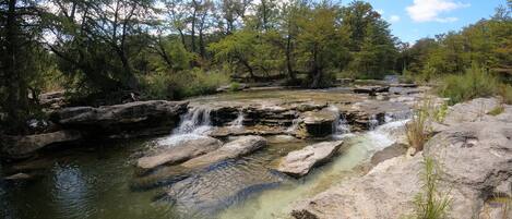 A natural landscape with a clear stream flowing over rocks, surrounded by trees and greenery.