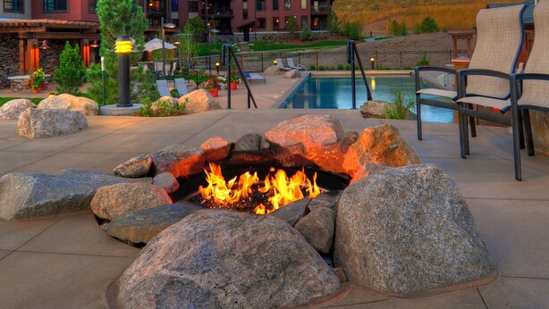 Una fogata rodeada de rocas grandes, una piscina, sillas de descanso y un edificio al fondo.
