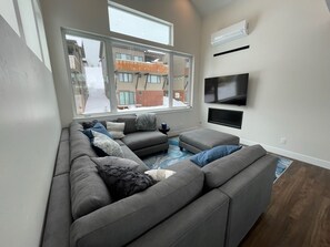 Un salon moderne avec un canapé d’angle gris, un téléviseur à écran plat fixé au mur et une grande fenêtre donnant sur l’extérieur.