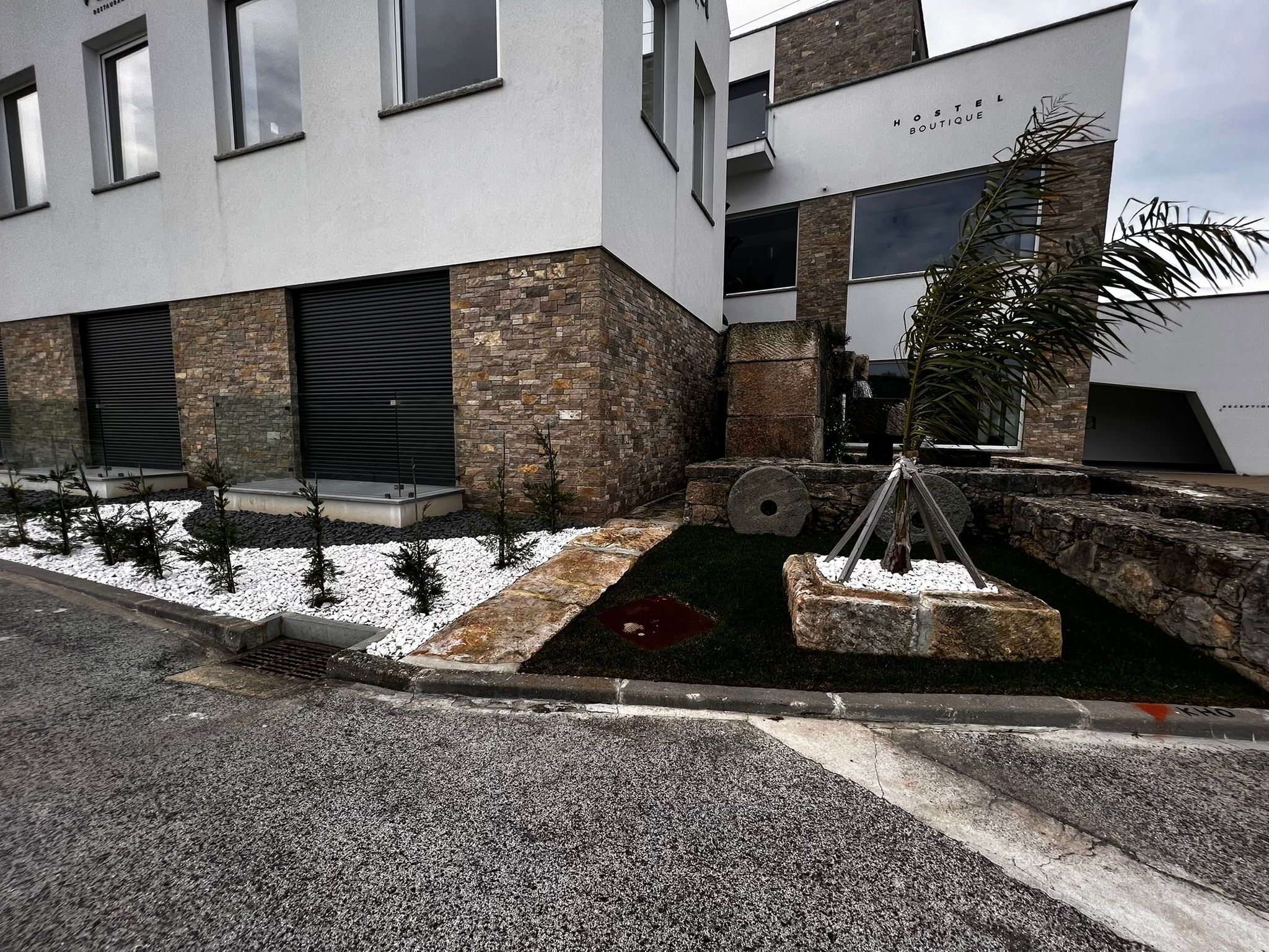 A modern building with a stone facade, a sign reading "HOTEL BOUTIQUE," and a landscaped garden area with small trees and a palm plant.