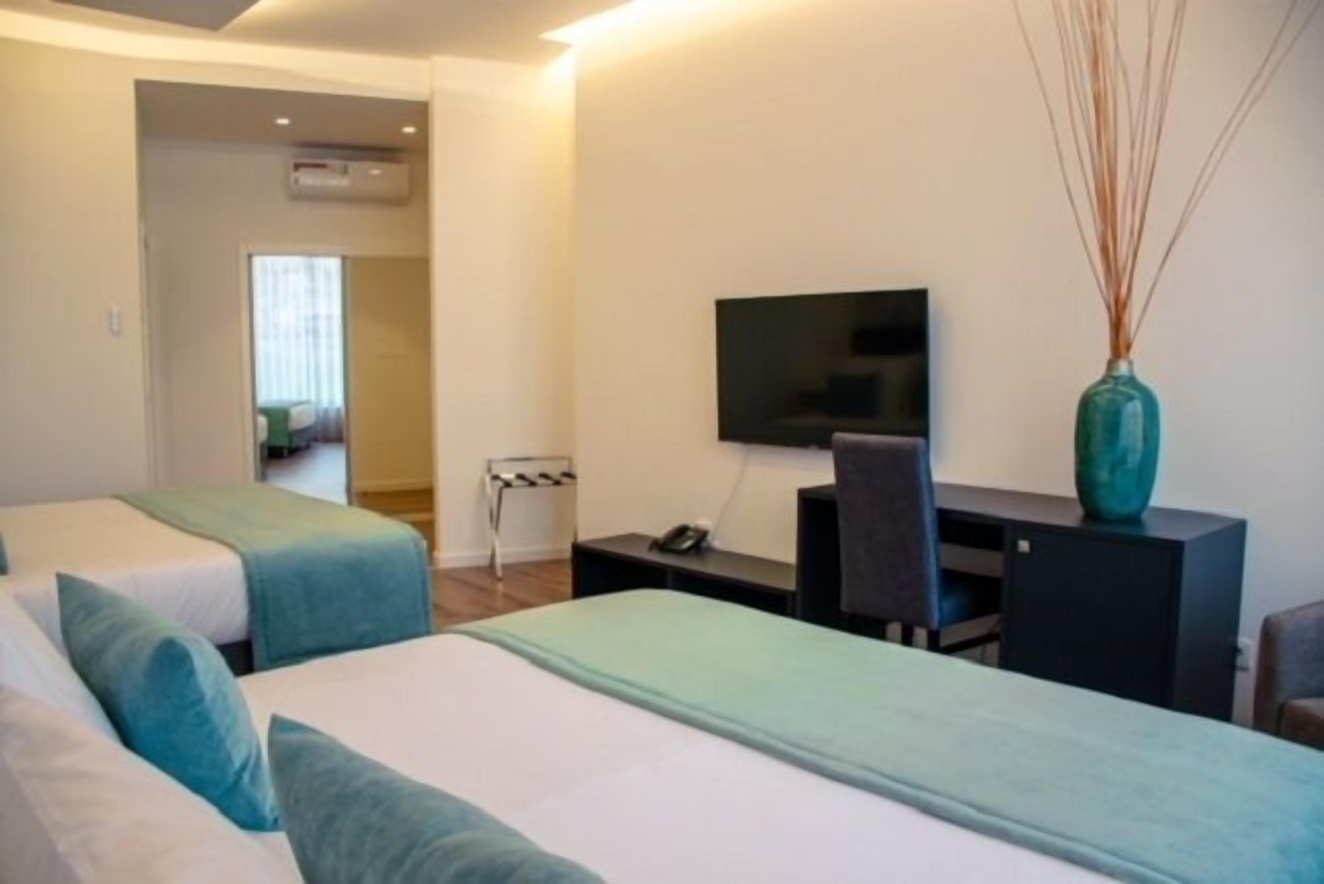 Quarto de hotel com duas camas, uma escrivaninha, uma cadeira, uma televisão e um vaso.