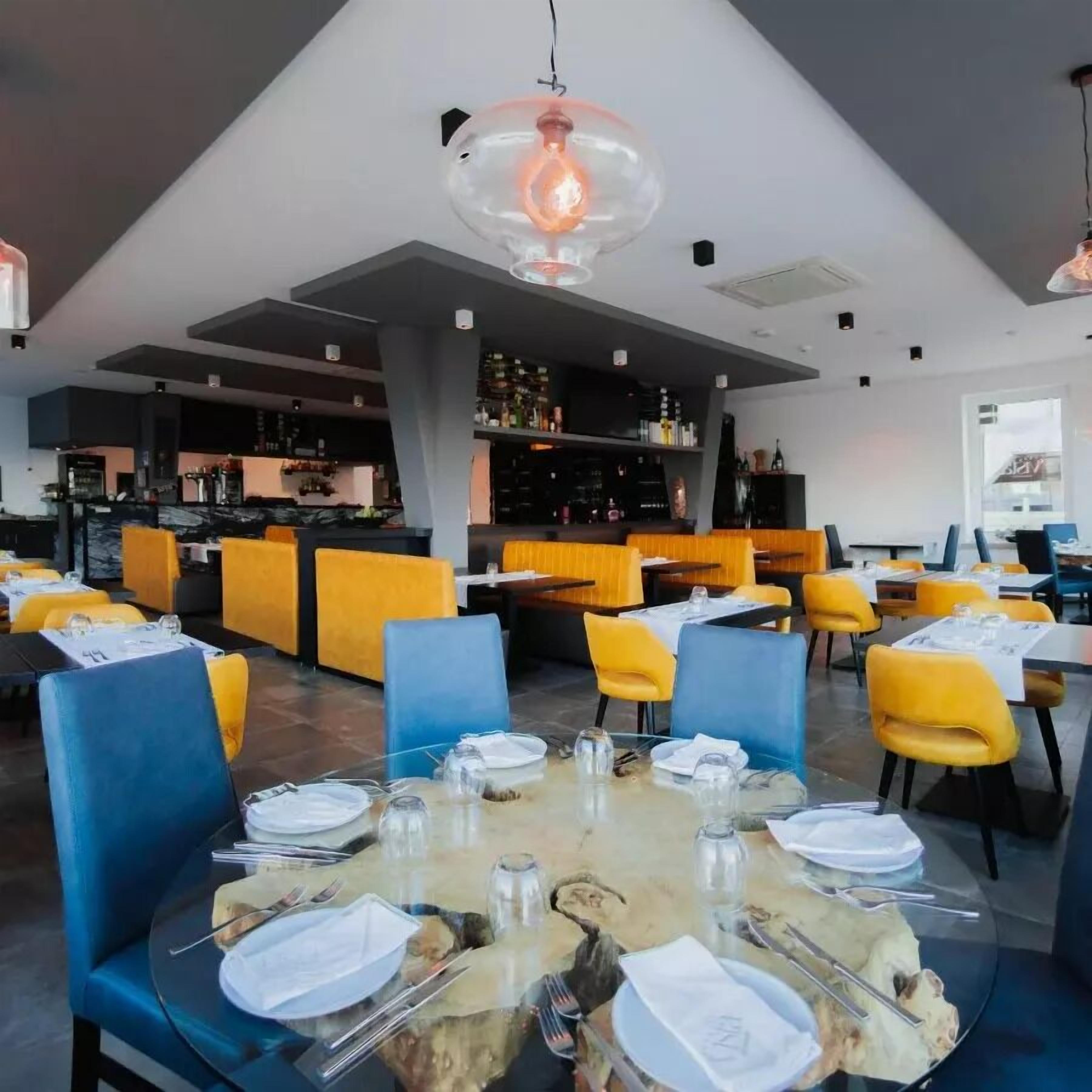 A restaurant with round tables, yellow and blue chairs, and a bar area in the background.