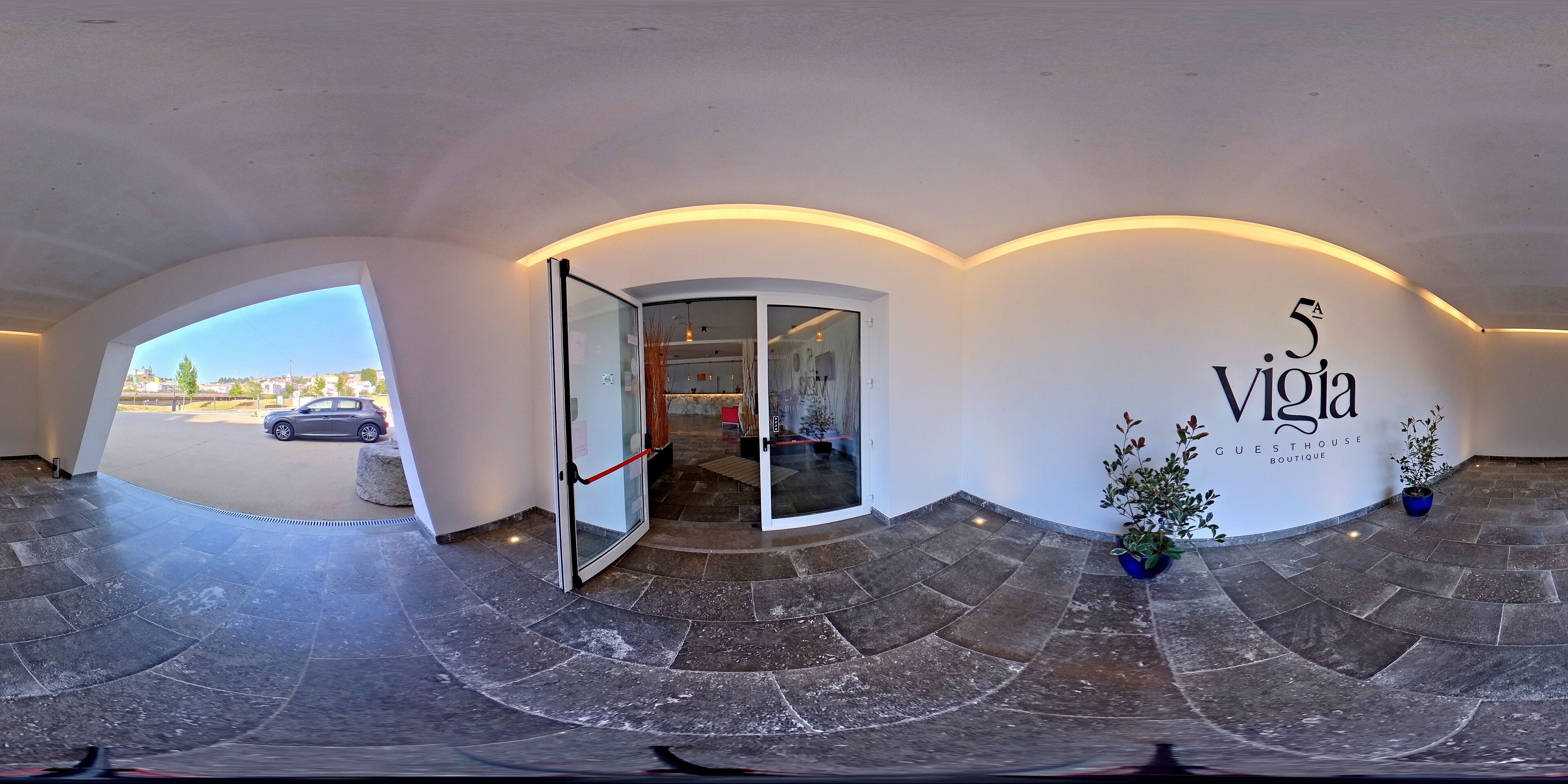 A 360-degree view of a hotel lobby with a glass door entrance, a stone-tiled floor, and a wall with the hotel's name and "GUESTHOUSE BOUTIQUE" sign.