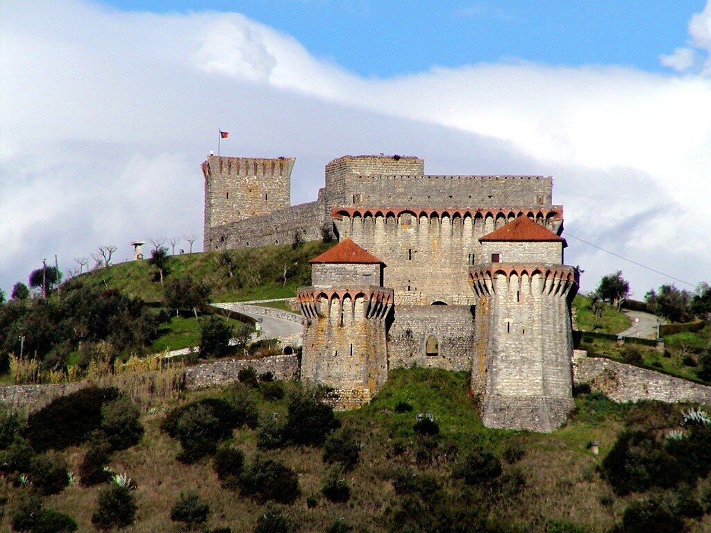 A historic stone castle with multiple towers and a flag on top, surrounded by greenery and hills.