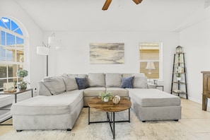 Un salon moderne avec un canapé d’angle, une table basse ronde et une grande fenêtre équipée de stores.