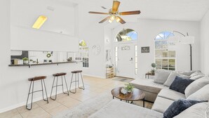 A spacious living area with a large sofa, a coffee table, and a ceiling fan. There is a kitchen area with a countertop and stools. - Snowbird Retreat! Escape Winter and Save Today (Panama City Beach)