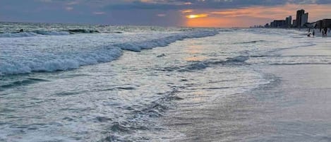 Una playa con olas, un atardecer y un horizonte lejano con el perfil de una ciudad.