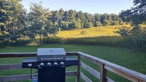A barbecue grill on a wooden deck with a grassy field and trees in the background. - Cozy Log Cabin at Warrior Ethos Farm (Cooperstown)