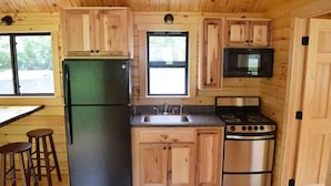 A compact kitchen with wooden cabinets, a refrigerator, a stove, a microwave, and two stools. - Cozy Log Cabin at Warrior Ethos Farm (Cooperstown)