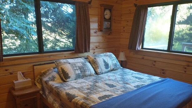 A wooden cabin bedroom with a bed, a nightstand, a clock, and large windows. - Cozy Log Cabin at Warrior Ethos Farm (Cooperstown)
