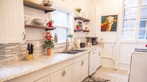 A modern kitchen with white cabinets, granite countertops, and a stainless steel sink. There are shelves above the counter holding various kitchen items and a painting on the wall. - The Kentucky Rose - Old Louisville with deck and courtyard (Louisville)