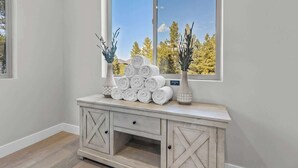 Un coin agrémenté d’une console blanche où sont posés des serviettes pliées et des vases décoratifs, ainsi qu’une grande fenêtre offrant une vue sur les arbres et un ciel dégagé.