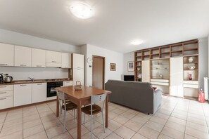 A modern kitchen with white cabinets, a dining table with chairs, a gray sofa, and a built-in shelving unit with decorative items.