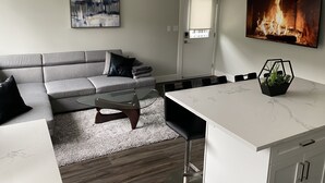 Una sala moderna con un sofá gris, una mesa de centro de vidrio y una isla de cocina de mármol blanco con una planta.