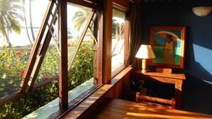 Una habitación con un banco de madera, una mesa, una lámpara y un cuadro. Se ve un ventanal con vistas a palmeras y flores.